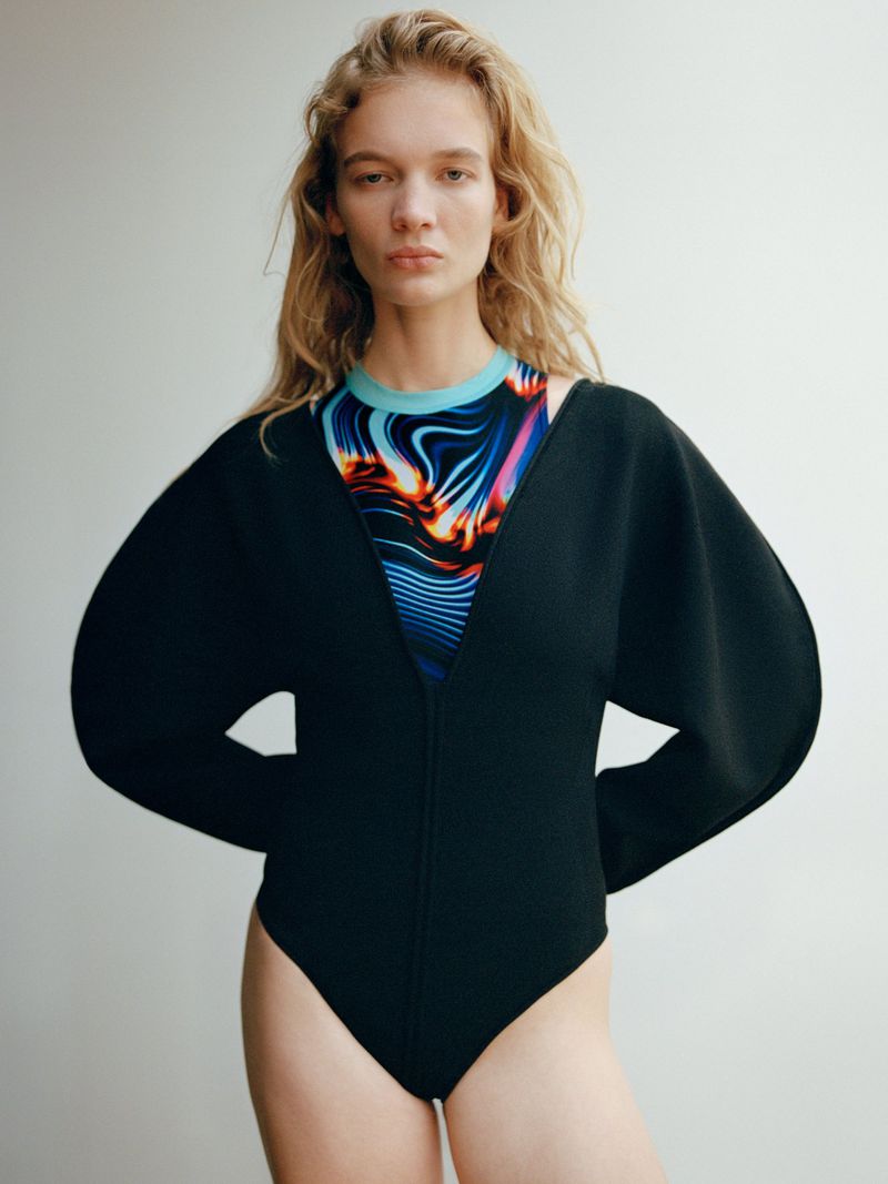 A fair-skinned woman with curly blonde hair, wearing a black bodysuit with wide sleeves, stands in front of a light grey backdrop.
