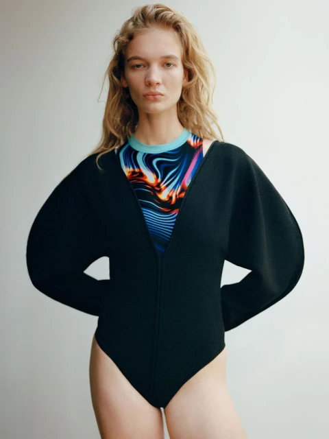 A fair-skinned woman with curly blonde hair, wearing a black bodysuit with wide sleeves, stands in front of a light grey backdrop.