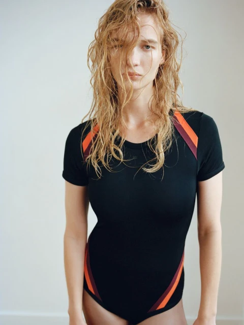 Wet-haired adult female fashion model in a black bodysuit poses in a brightly lit, neutral-toned photo studio against a light-colored background.