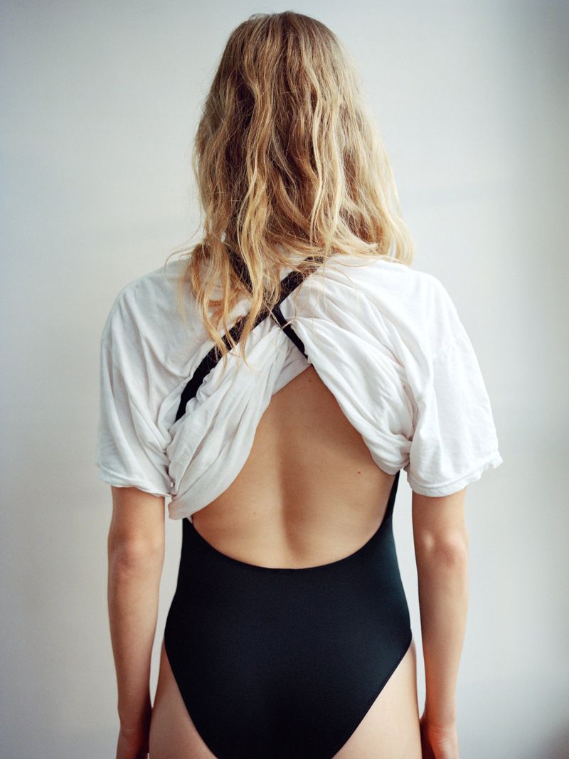 Back view of a fair-skinned woman with blonde hair wearing a white T-shirt and a black one-piece swimsuit against a neutral backdrop.