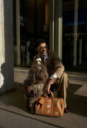 A man in a tan suit kneeling beside a curly brown dog holding a leather duffel bag in front of a building exterior.