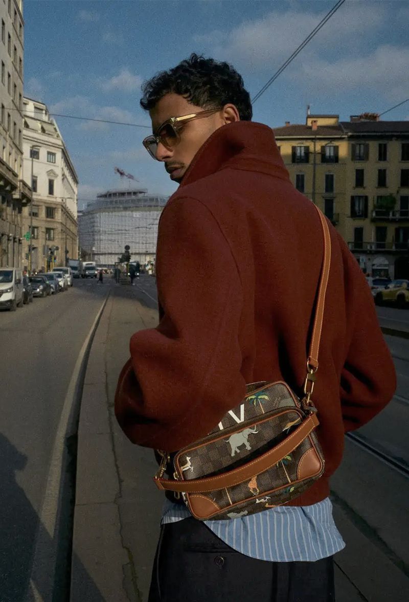 Stylish adult male wearing sunglasses, a maroon jacket, and carrying a patterned bag walks down a city street on a sunny day.