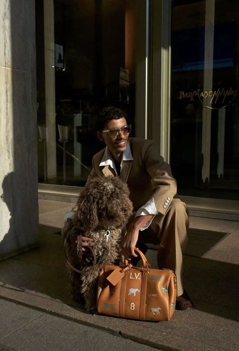 Stylish man with sunglasses and a tan suit poses outdoors with his dog and a designer duffel bag on a sunny day in an urban setting.