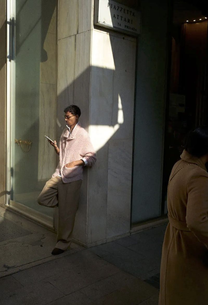 Woman in pink jacket leaning against building in dappled light while looking at her phone, with another person walking by.