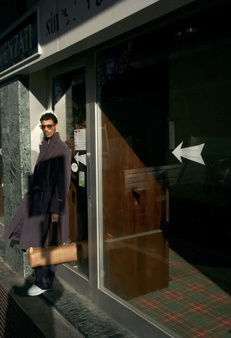 Model wearing a dark coat and holding a tan leather duffel bag while standing outside a storefront with strong sunlight and shadows.