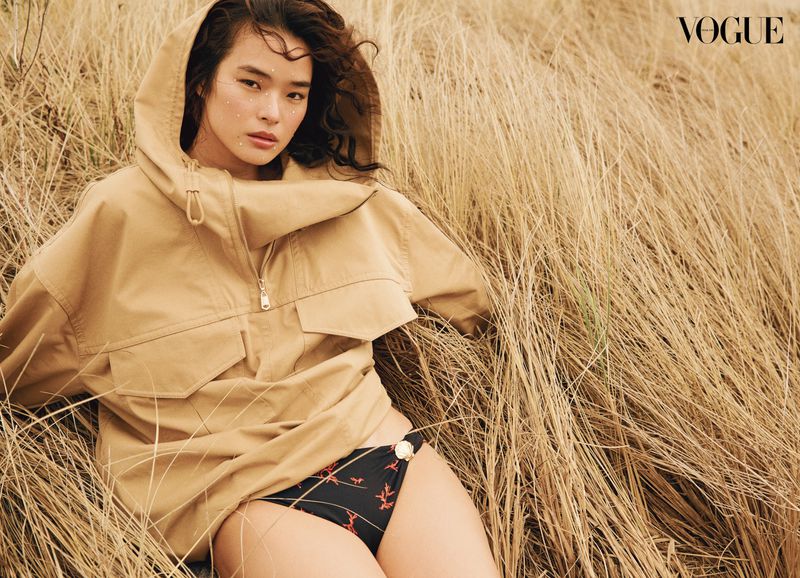 Asian woman in oversized tan hooded jacket and black bikini bottoms lying in field of tall, dry grass in outdoor fashion shot.