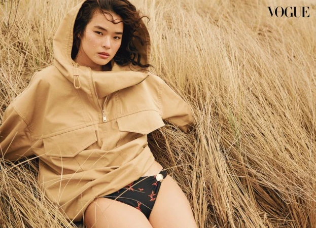 Asian woman in oversized tan hooded jacket and black bikini bottoms lying in field of tall, dry grass in outdoor fashion shot.