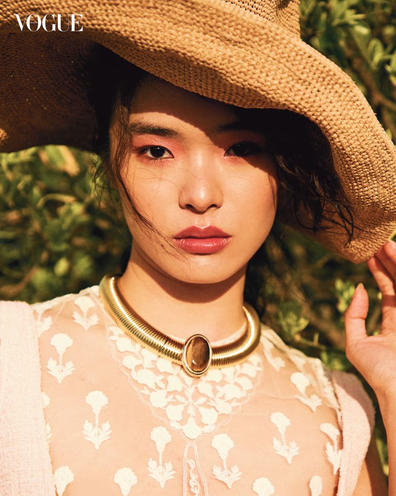Close-up of a young Asian woman wearing a straw hat and statement necklace outdoors in front of a green leafy background. Vogue logo is visible.