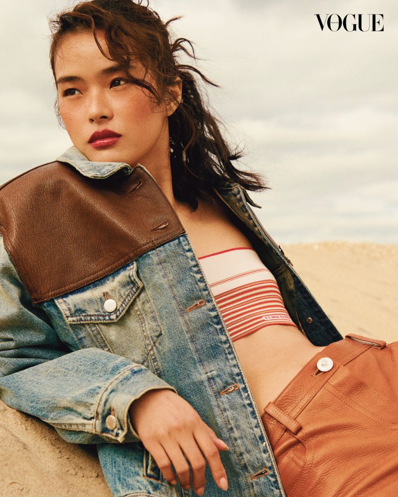 Attractive Asian woman in stylish denim jacket, tube top and leather pants posing outdoors against a sandy, cloudy background.