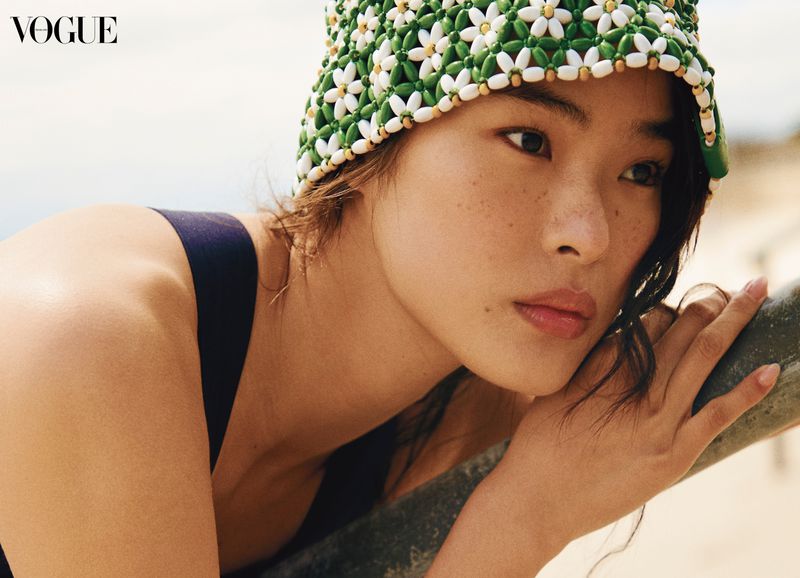 Portrait of an Asian woman wearing a green and white beaded hat, black top, and soft makeup in a close-up Vogue fashion editorial.