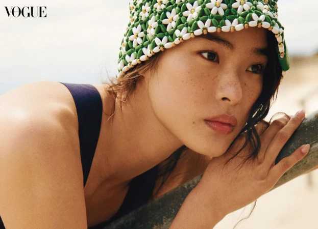Portrait of an Asian woman wearing a green and white beaded hat, black top, and soft makeup in a close-up Vogue fashion editorial.