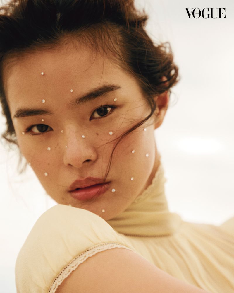 Portrait of a young Asian woman with jeweled makeup, wearing a light yellow dress in front of a light background. Vogue logo visible.