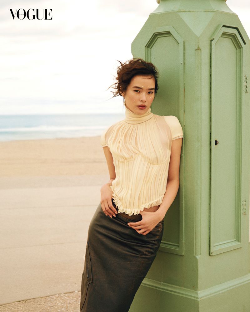 Fashion editorial shot of a woman in a light-yellow top and dark-brown skirt, leaning against a green post on a beach location.