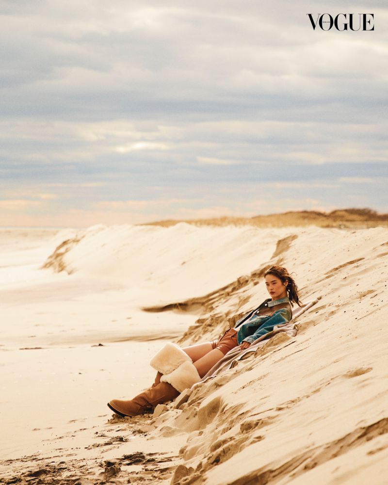 A young woman in a blue jacket and boots poses lying on a sandy beach on a cloudy day, with the Vogue logo visible in the upper corner.