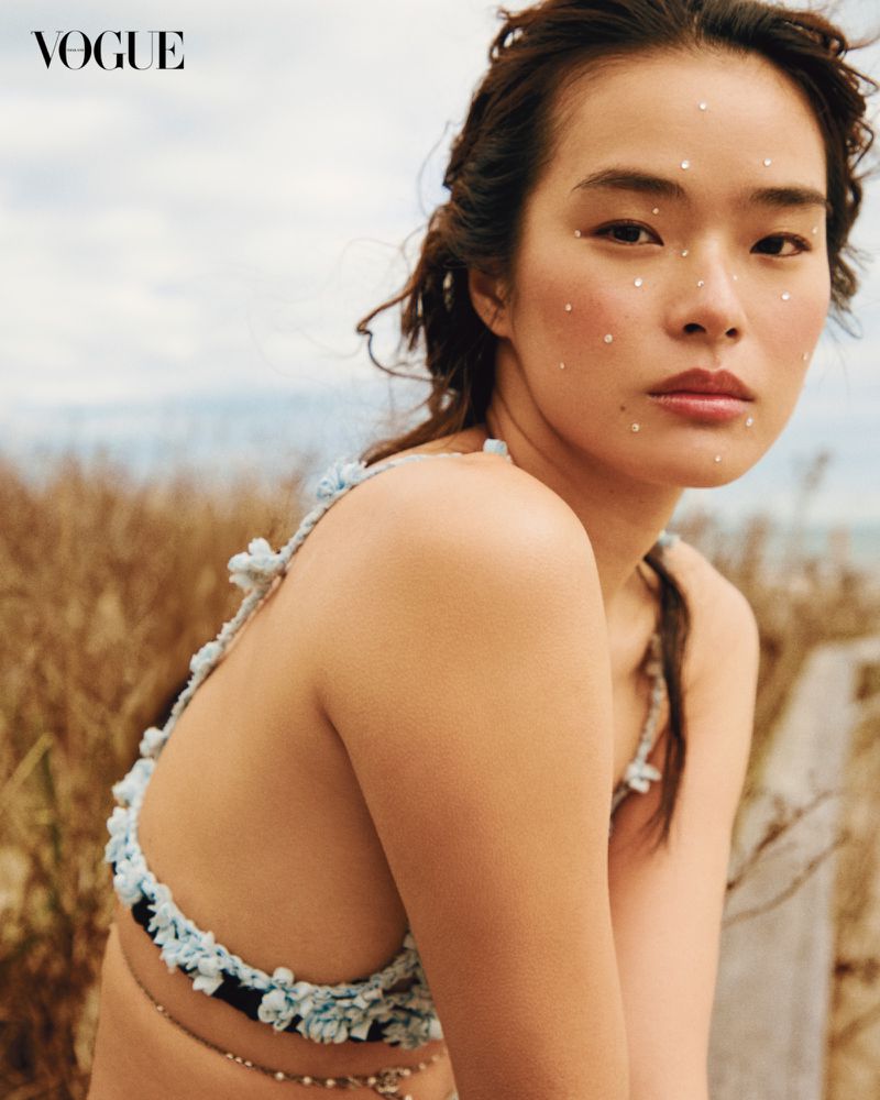 Close-up shot of a young Asian woman with pearls on her face, wearing a floral bikini top against a natural outdoor backdrop for Vogue.