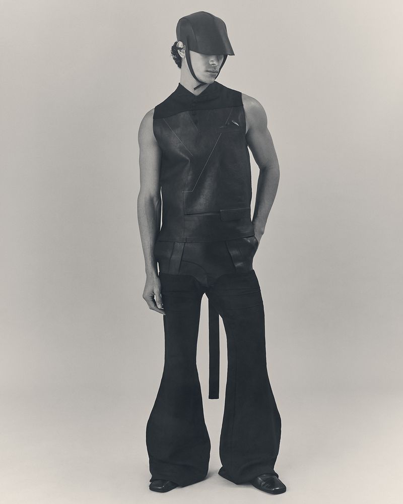 Full-length studio portrait of a person wearing black vest, wide-leg pants, and cap against a neutral background, in a fashion editorial.