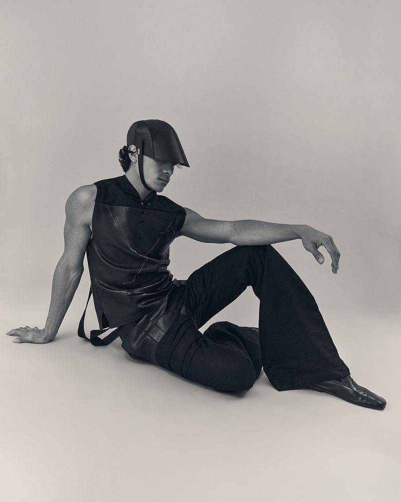 Stylish man in black sleeveless top, loose pants, cap sitting indoors. Soft lighting emphasizes fashion, posing, and studio setting.