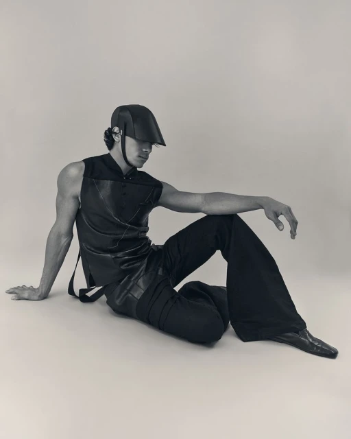 Stylish man in black sleeveless top, loose pants, cap sitting indoors. Soft lighting emphasizes fashion, posing, and studio setting.