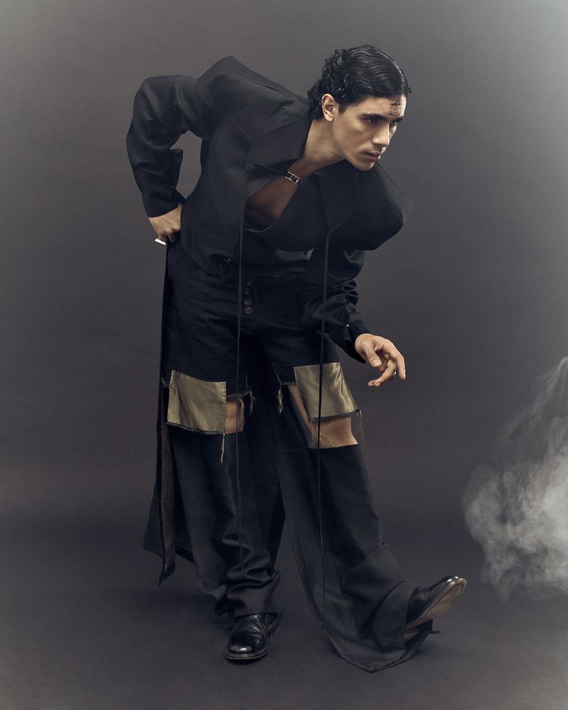 Man in an all-black fashion ensemble poses in a studio, angled stance, showcasing deconstructed details against a plain backdrop with smokey ambiance.