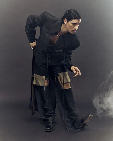 Man in an all-black fashion ensemble poses in a studio, angled stance, showcasing deconstructed details against a plain backdrop with smokey ambiance.