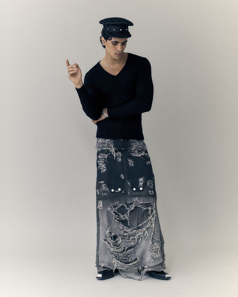 A fashion model in a black sweater and deconstructed denim maxi skirt poses in front of an off-white backdrop in a studio shot.
