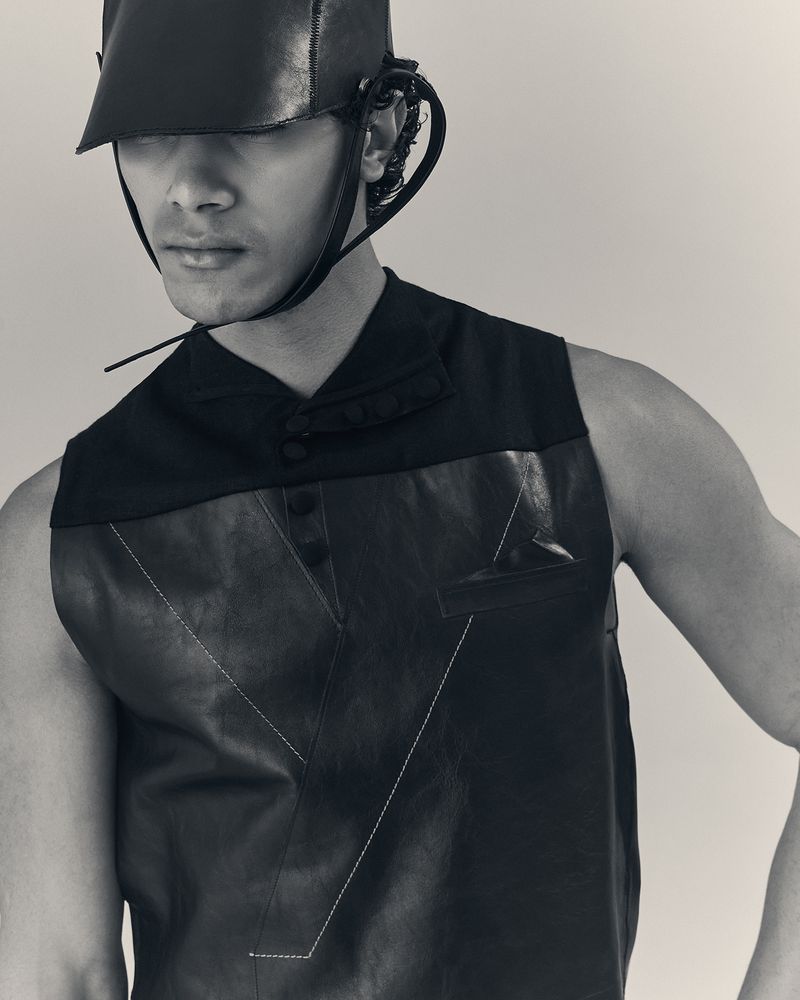 Monochrome portrait of a male model wearing a black leather vest and futuristic headwear in a studio setting with soft, diffused lighting.