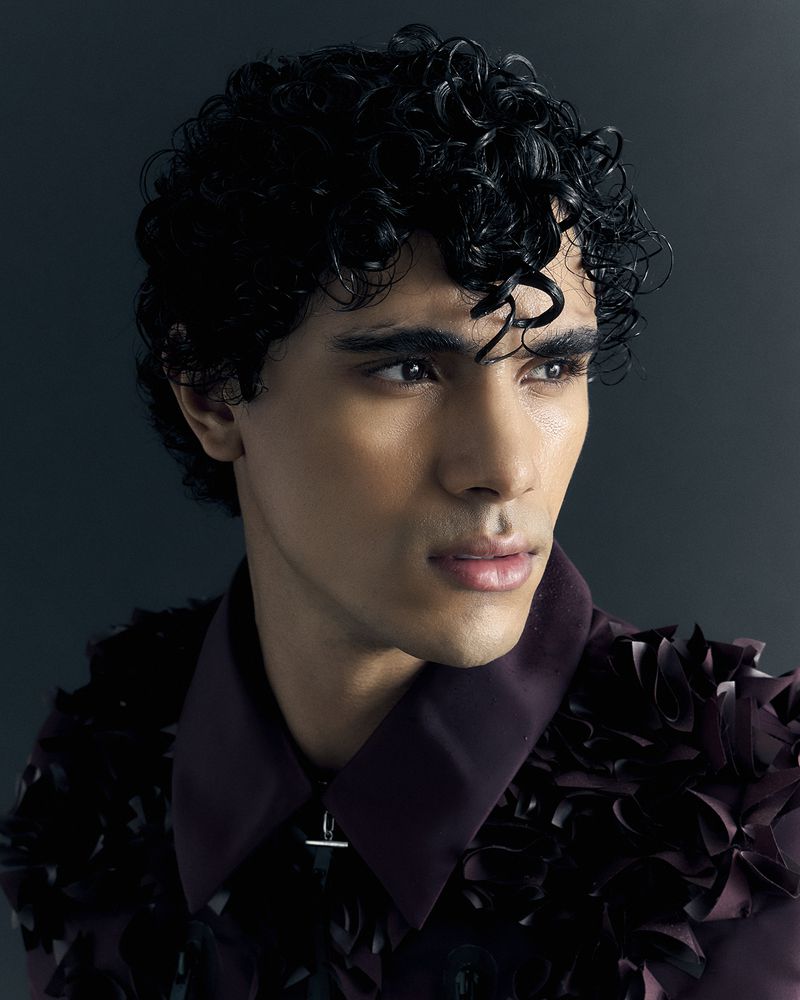 Portrait of a dark-haired male model with curly hair wearing a plum-colored jacket with decorative embellishments against a dark background.