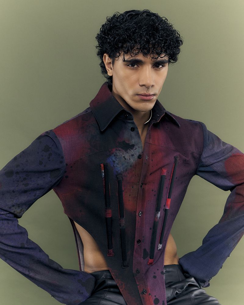 Portrait of a curly-haired male model in a vibrant, patterned shirt posing indoors against an olive-green backdrop.