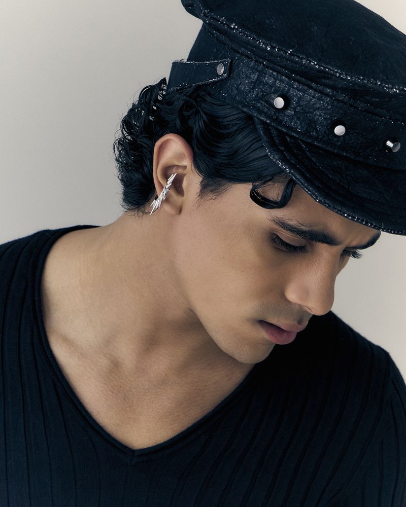 Close-up of a young man with an earring and a black cap, wearing a black v-neck top, against a neutral background in portrait orientation.