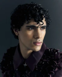 Close-up portrait of a male model with dark curly hair wearing a dark plum textured shirt against a dark grey background.