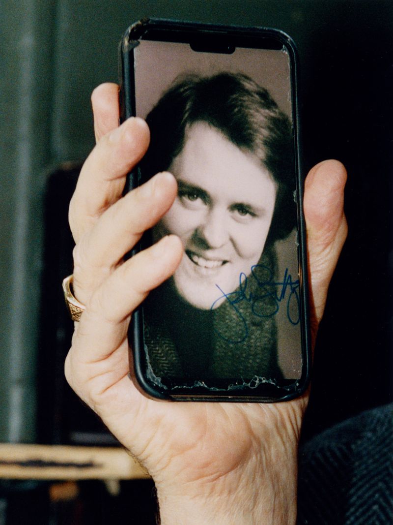 Hand holds a smartphone displaying a smiling person's photograph with a signature on the screen, set against a dark background.