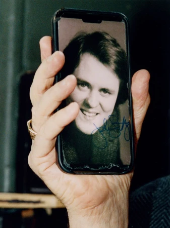 Hand holds a smartphone displaying a smiling person's photograph with a signature on the screen, set against a dark background.