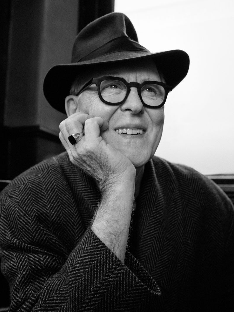 Black and white portrait of an elderly man wearing a hat and glasses, resting his chin on his hand with a smile. He is wearing a herringbone jacket.
