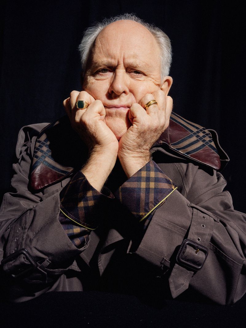 Portrait of an elderly man with grey hair wearing a brown plaid trench coat, posing with his hands under his chin in a dark studio.