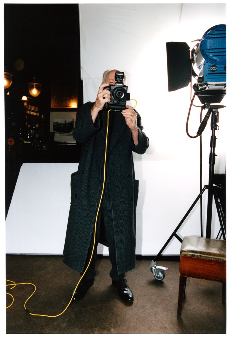 A man in a dark coat holds a camera in a photo studio with lighting equipment. A yellow cable extends from the camera to the floor.