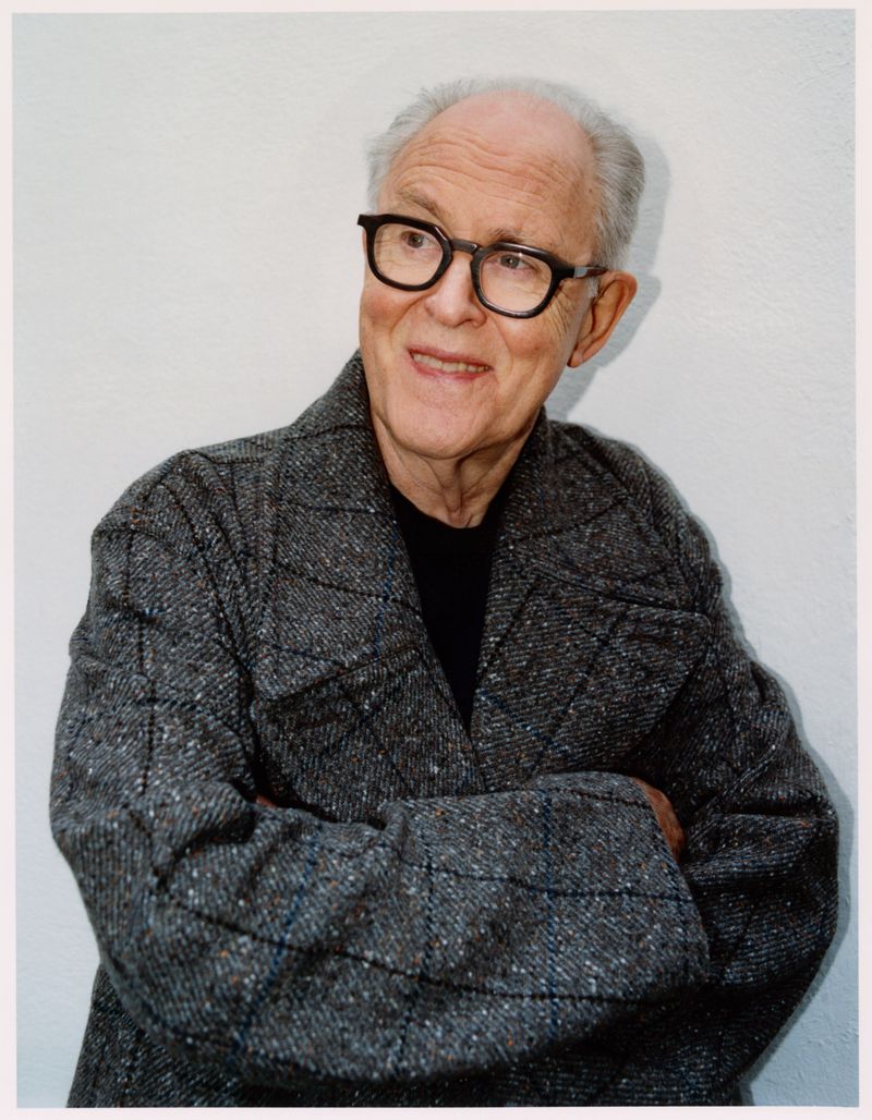 Elderly man with glasses and grey tweed coat, arms crossed, smiling in a simple indoor setting. Upper body portrait with soft lighting.