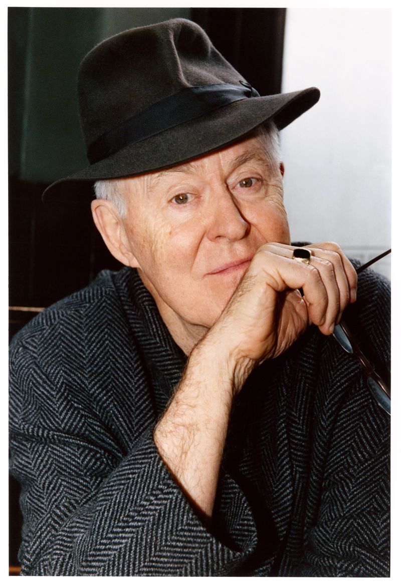 Portrait of an older man with a hat, wearing a patterned coat, looking thoughtfully into the distance with his hand to his face.