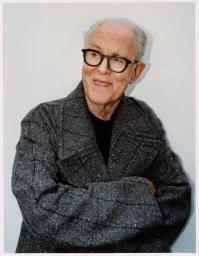 John Lithgow wearing a textured patterned coat and black framed glasses against a white studio background.