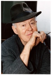 Portrait of actor John Lithgow wearing a dark fedora and herringbone jacket, holding glasses near his face for American Vogue.
