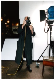 Man wearing a dark long coat holding a camera in front of his face in a studio with a bright backdrop and lighting equipment