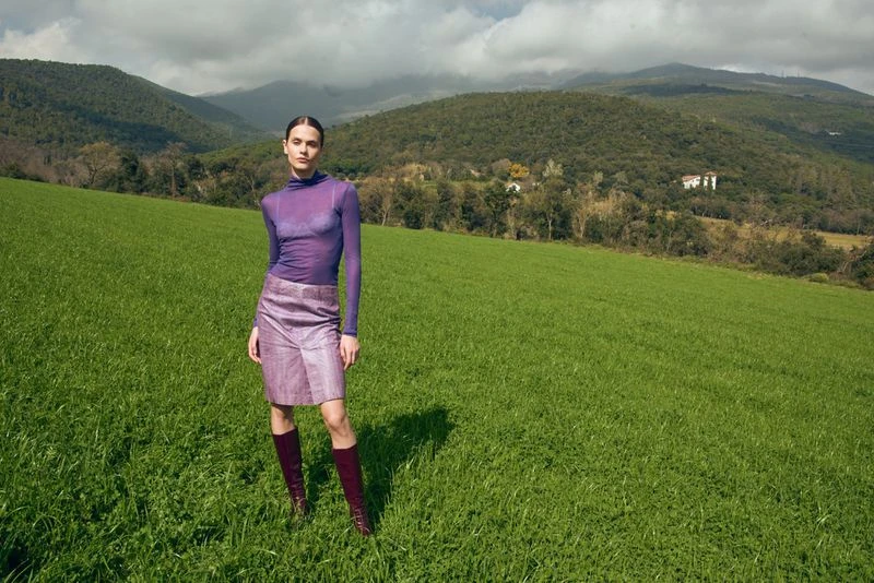Fashion shot of a woman in a purple mesh top, textured skirt, and maroon boots stands in a green field with mountains in the background.