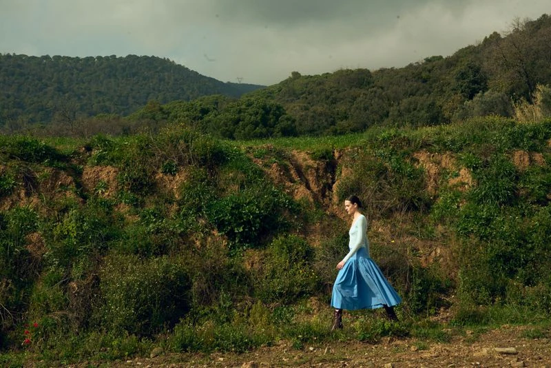 Adult woman in a blue dress walks on a dirt path surrounded by green grass and brush; a green mountain is visible in the background.