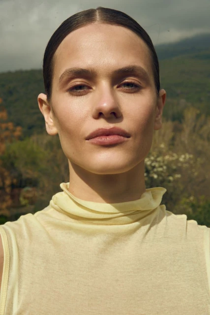 Close-up studio portrait of a woman with neutral expression, wearing a yellow top, with an outdoor background of trees and cloudy sky.