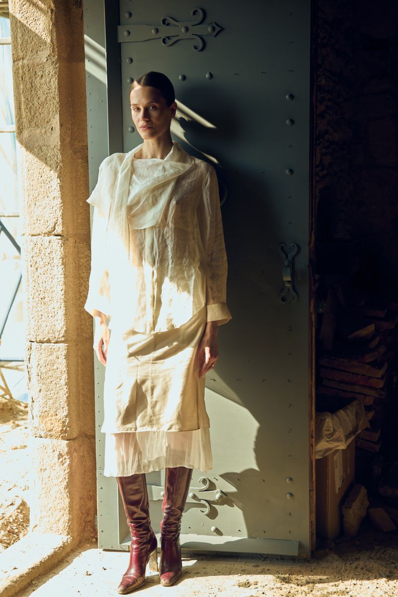 A woman in an off-white layered dress stands in a doorway with a blue door, light from the window on the left illuminates the scene.