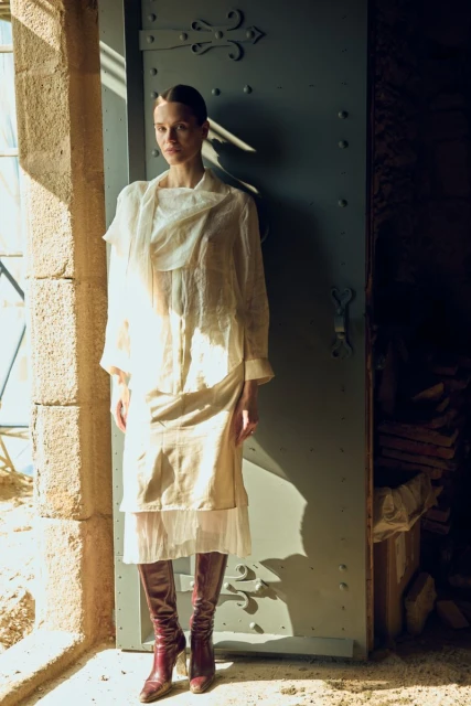 A woman in an off-white layered dress stands in a doorway with a blue door, light from the window on the left illuminates the scene.