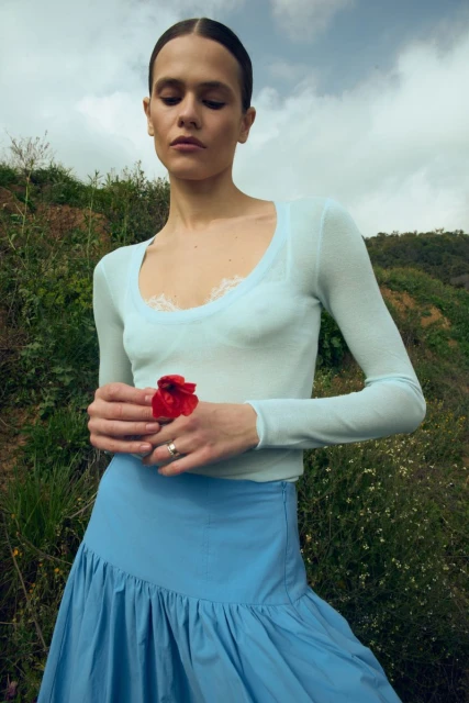 Woman in blue top and skirt holds red flower against a hillside backdrop with a cloudy sky, creating a serene, fashion-focused image.