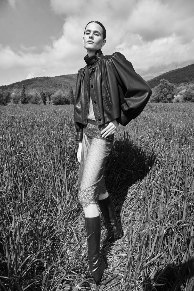 A woman in a black leather jacket and patterned skirt poses in a field of tall grass, with rolling hills and a cloudy sky in the background.