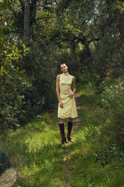 A woman in a yellow dress and dark brown boots stands on a grassy path in a sun-dappled forest setting under a canopy of trees.