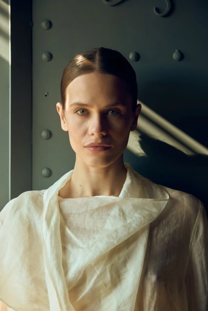 Close-up portrait of a woman with light skin and brown hair, wearing a light-colored top, posing in front of a metal door.