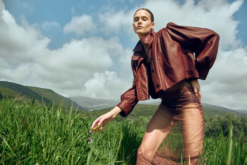 A fashion photo showcases a model in a brown leather jacket and sheer pants in a tall grassy field with a mountain background.